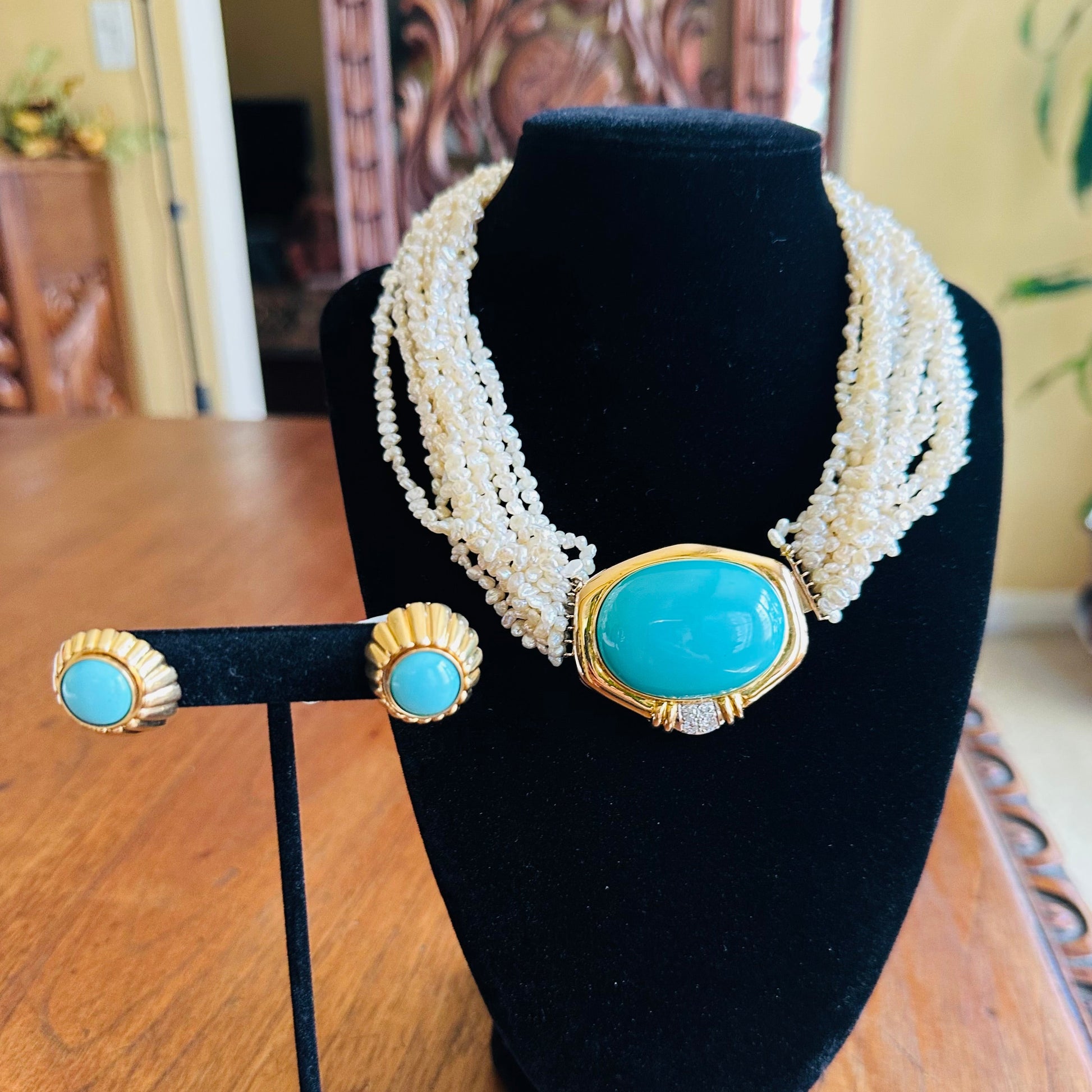 14k gold jewelry set displayed on black velvet stands, featuring a multi-strand pearl necklace with a large turquoise cabochon centerpiece and matching stud earrings with turquoise and gold scalloped settings, set on a wooden table.